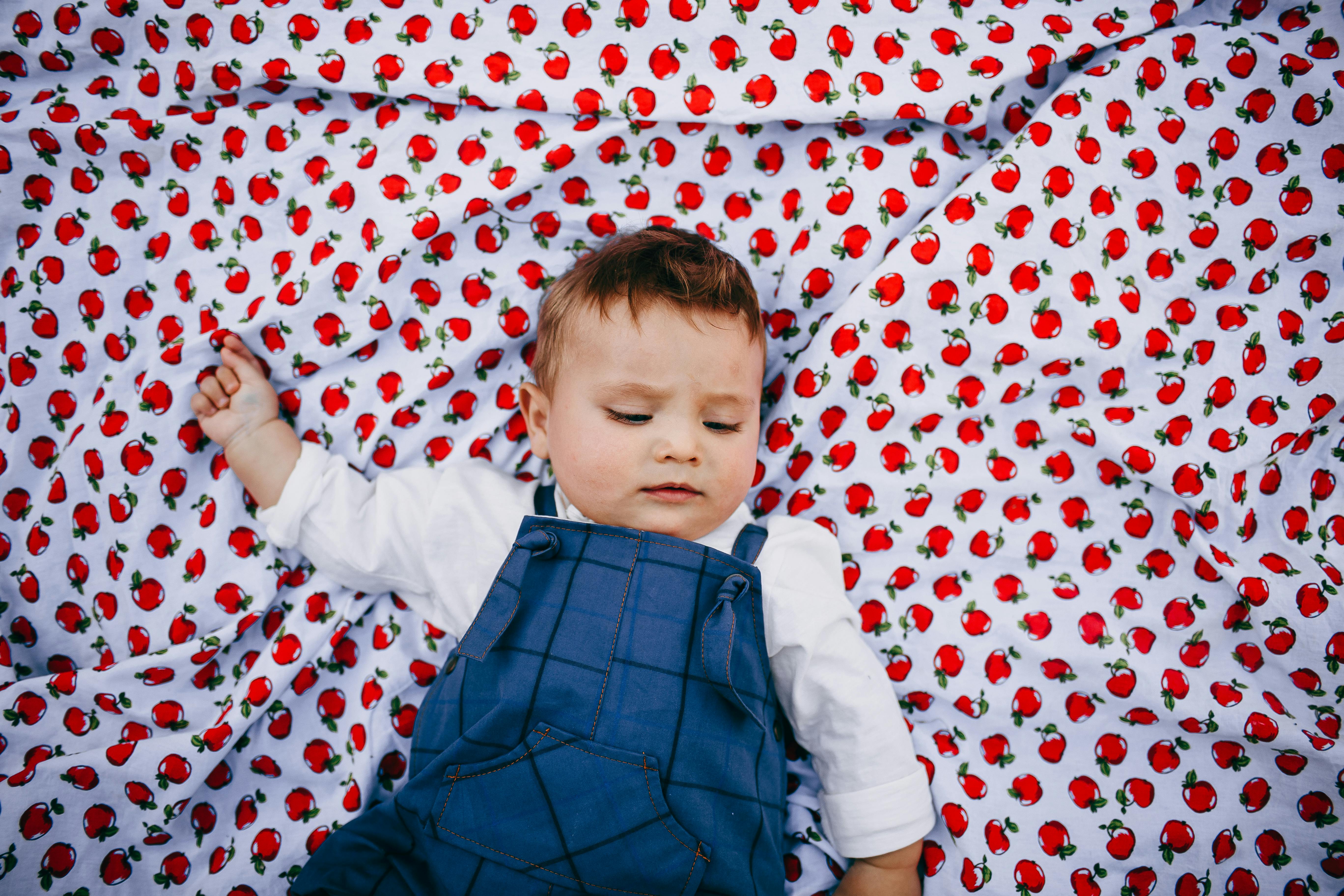 A cute baby in blue overalls resting on a vibrant strawberry print blanket.