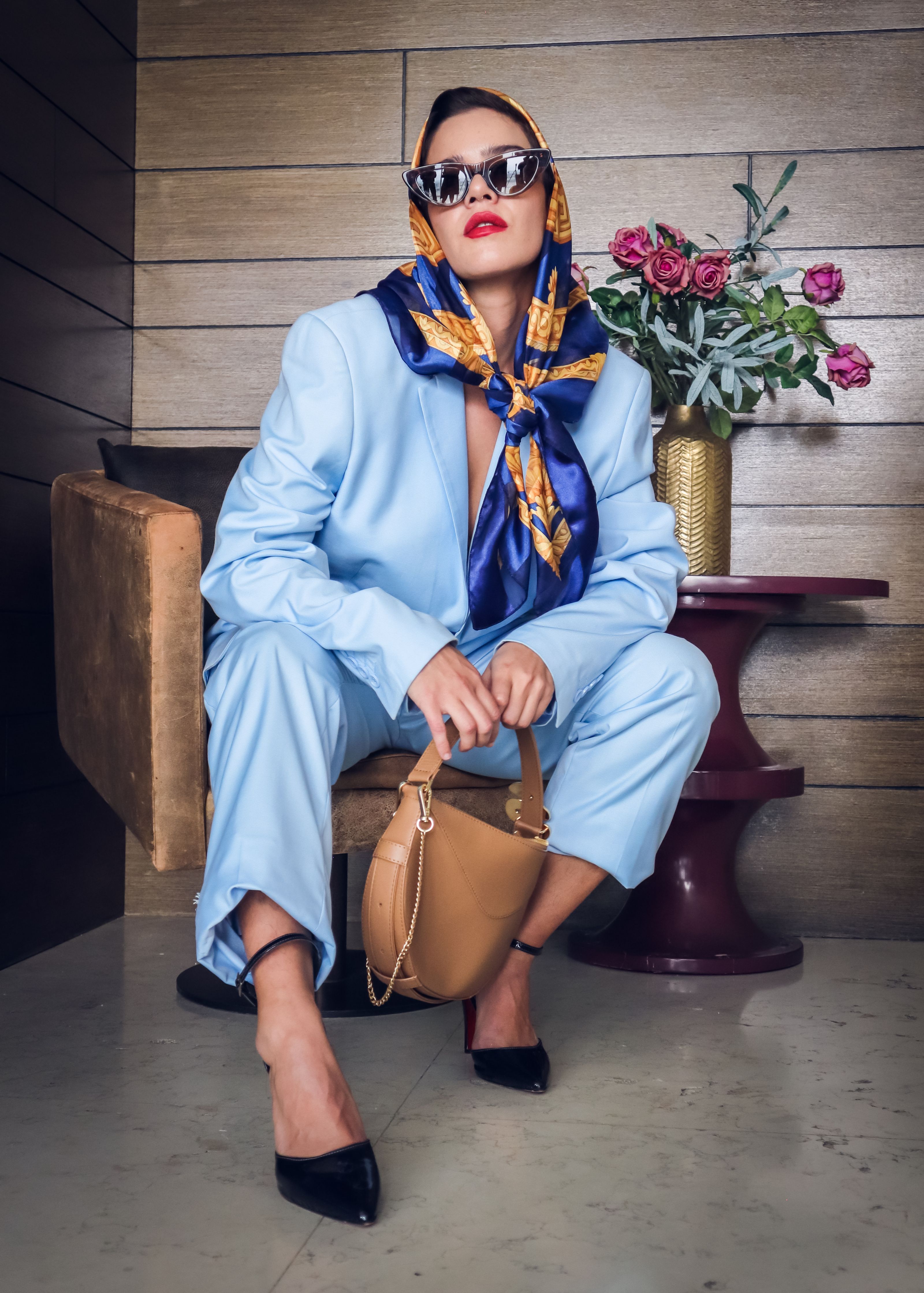 Elegante mulher em traje azul e lenço posando com óculos escuros e bolsa em ambiente interno.