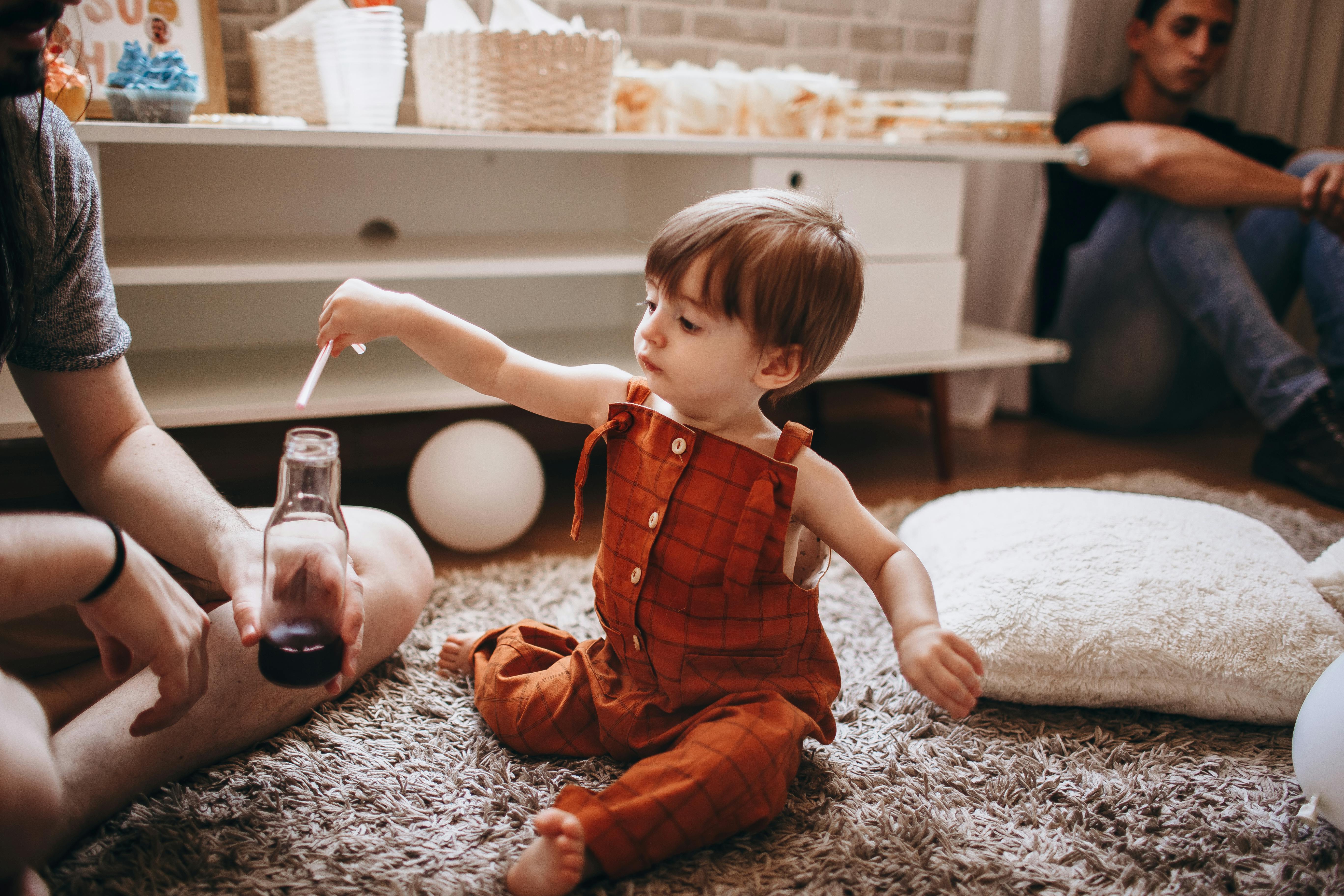 A toddler in an orange romper plays with a bottle indoors. Cozy family setting.