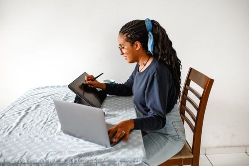 Free Young woman working on a digital tablet and laptop in a cozy home setting. Ideal for tech and lifestyle concepts. Stock Photo