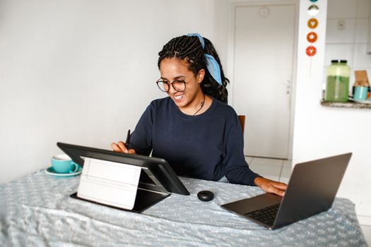 A young woman smiling while working on a laptop and drawing tablet at home.