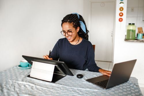 Free A young woman smiling while working on a laptop and drawing tablet at home. Stock Photo