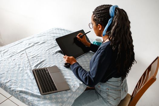 A young woman uses a drawing tablet and laptop in a home office setting.