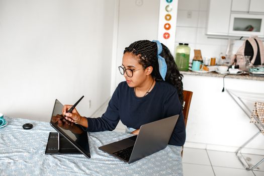 A young woman creatively works on a digital drawing tablet and laptop at a cozy home setup.