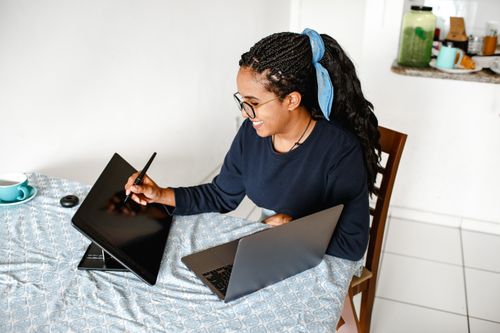 Free Smiling woman using digital tablet and laptop at home office, showcasing creativity and technology. Stock Photo