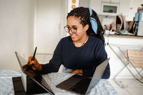 Free Smiling woman using tablet and laptop at home, epitomizing modern remote work. Stock Photo