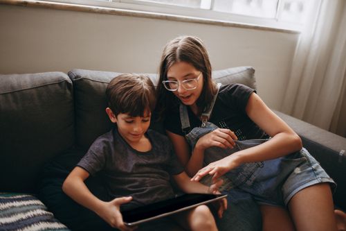 Free A teenage girl and a young boy happily using a tablet on a couch in a cozy living room setting. Stock Photo