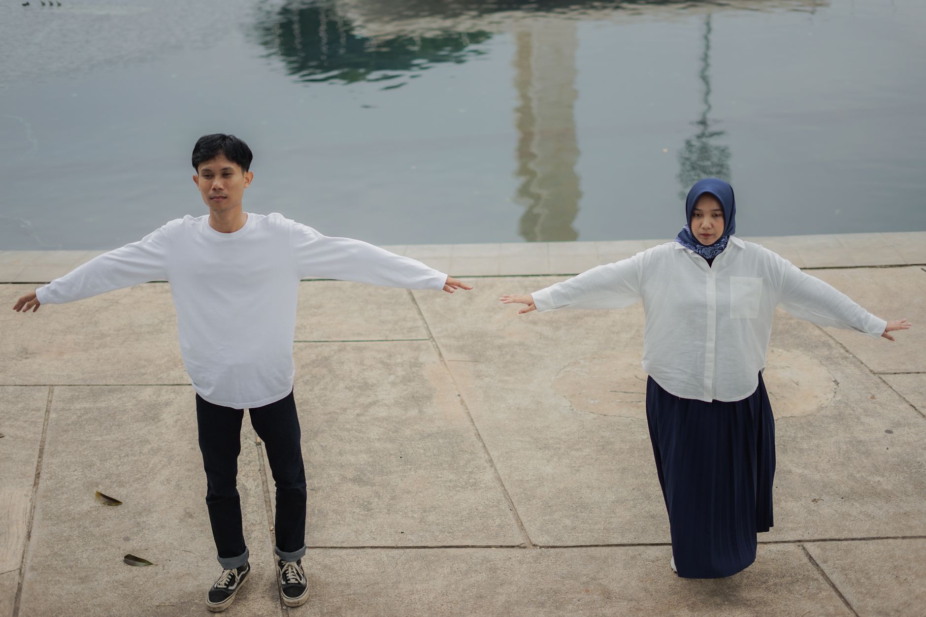 Two adults with outstretched arms meditating outdoors near tranquil water.