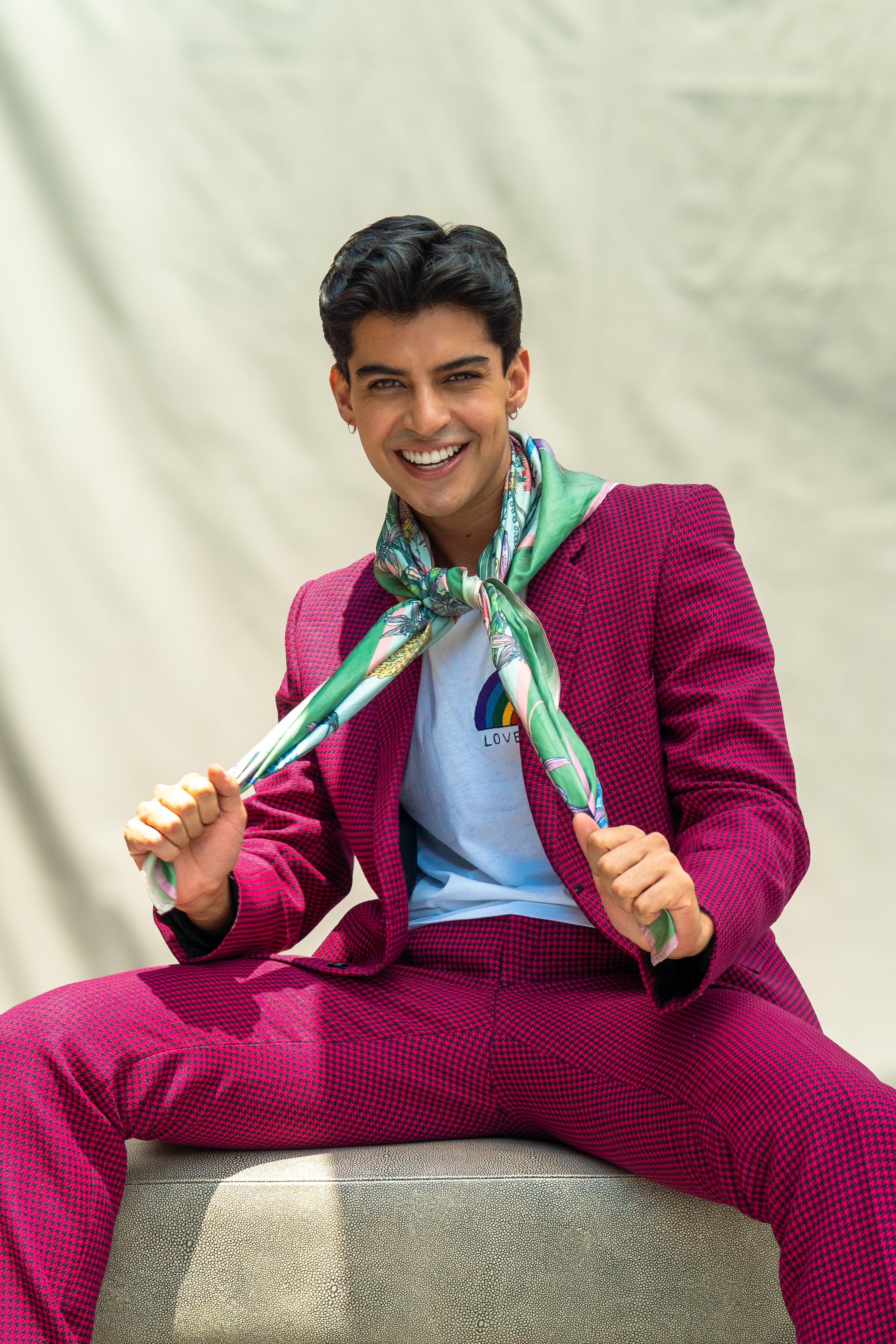 A young man in a pink suit and floral scarf smiles, embracing vibrant fashion outdoors.
