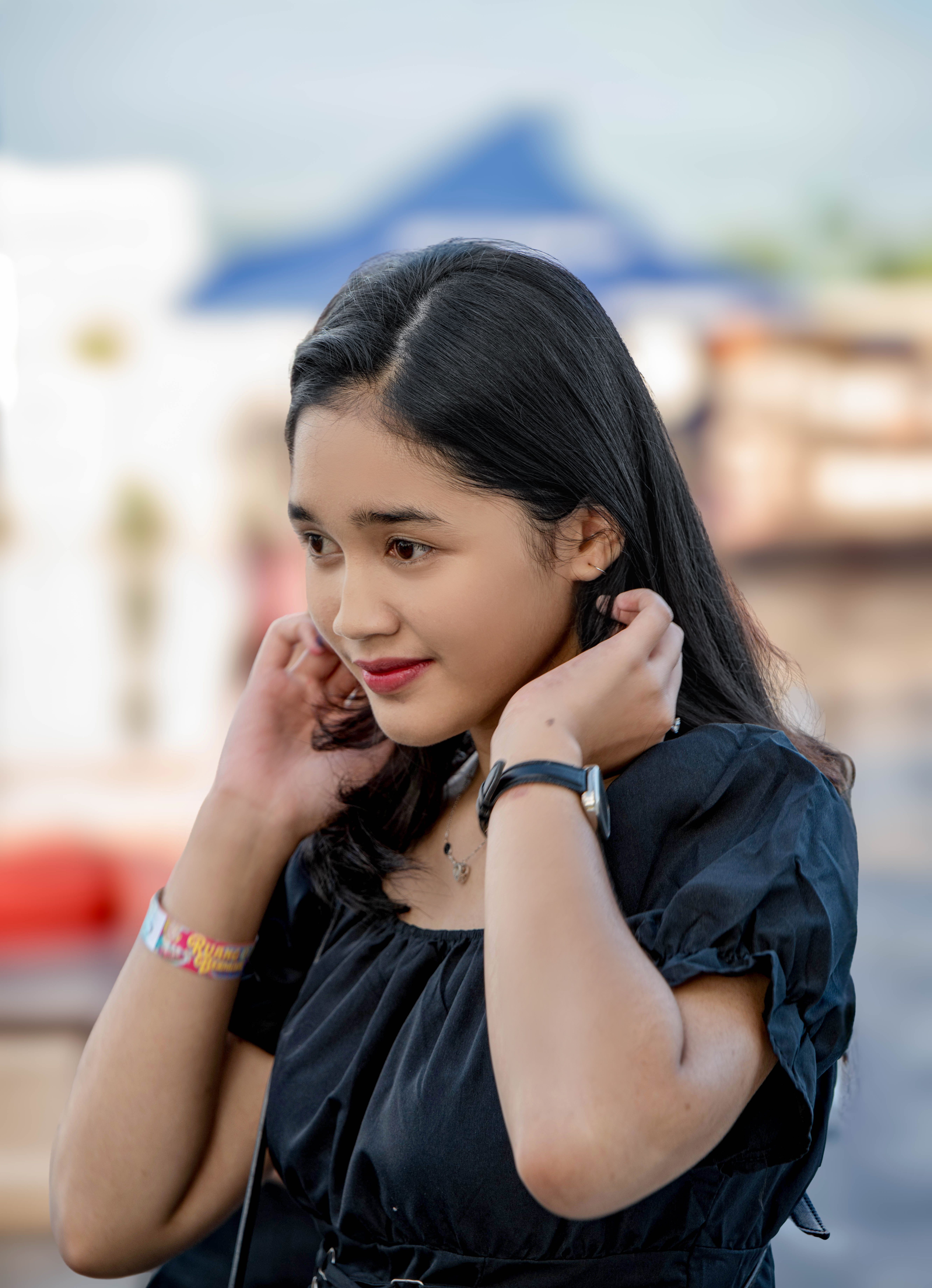 A young woman adjusts her hair outdoors, wearing a stylish black dress and colorful accessories.
