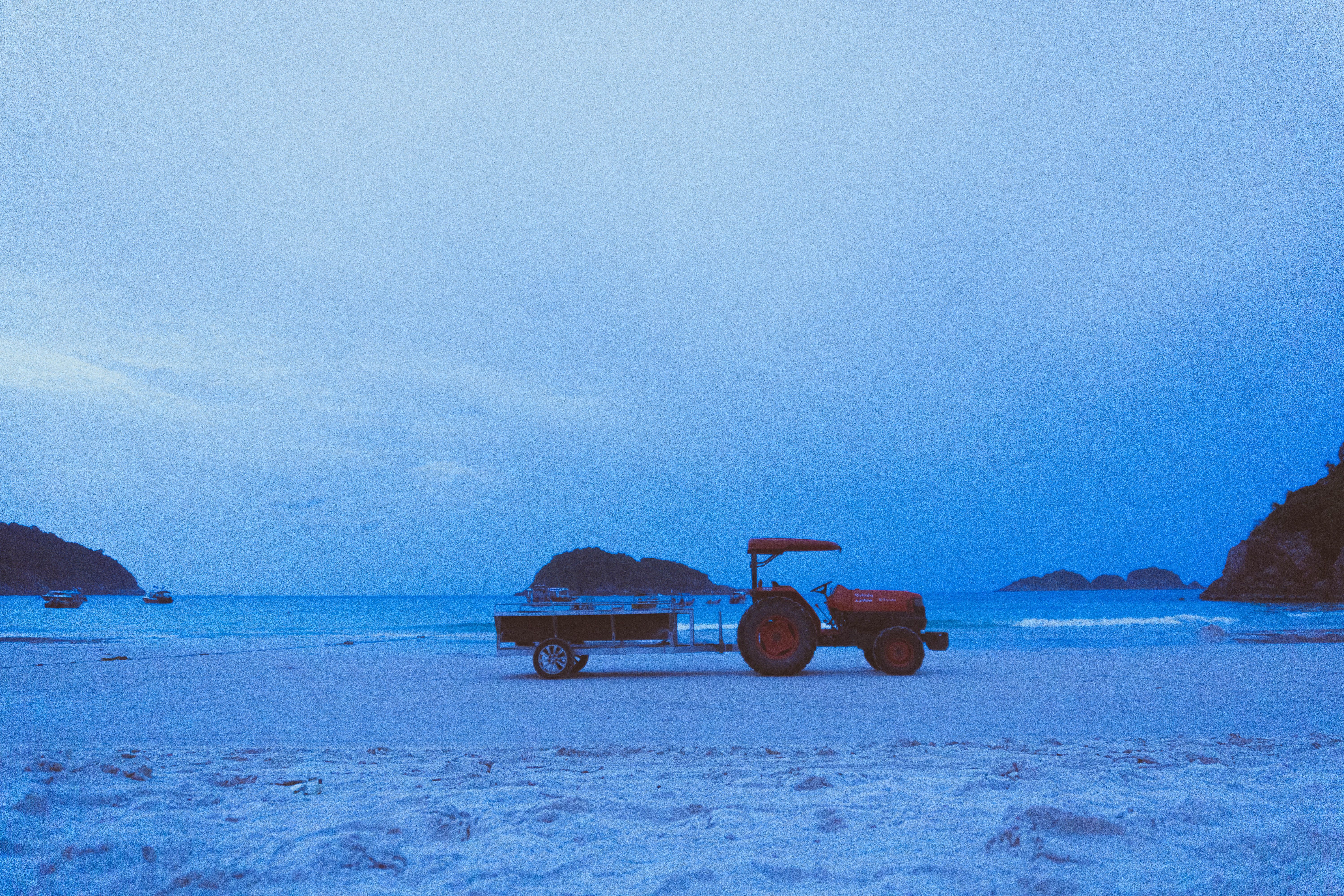 Tractor parked on a tranquil beach at dusk with an ocean view and distant islands under a blue sky.