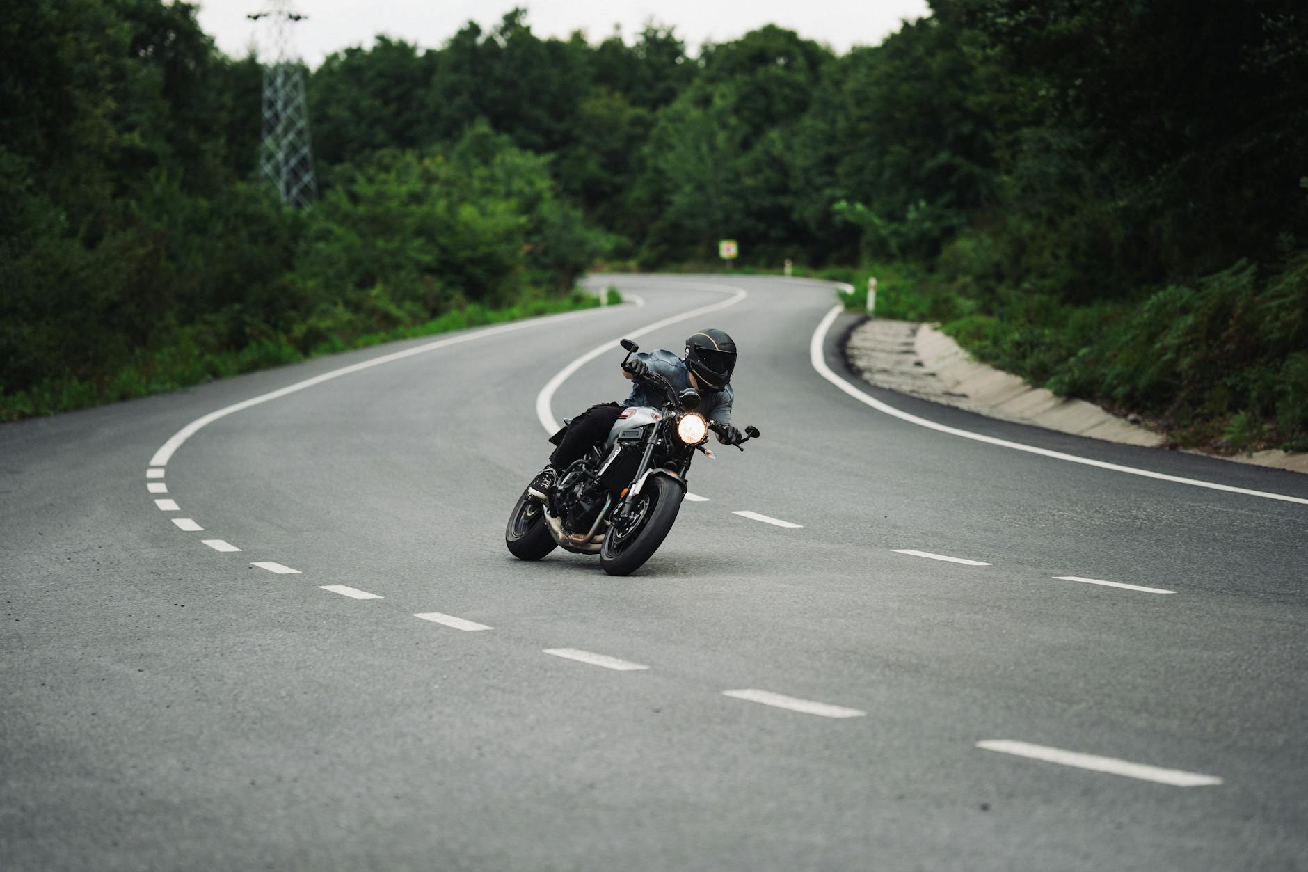 Pengendara motor dalam perlengkapan hitam yang mengemudikan tikungan di jalan indah yang dikelilingi oleh pepohonan hijau.
