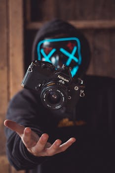 一个戴着发光面具的连帽人物悬浮在一台老式相机上方，营造出超现实的氛围