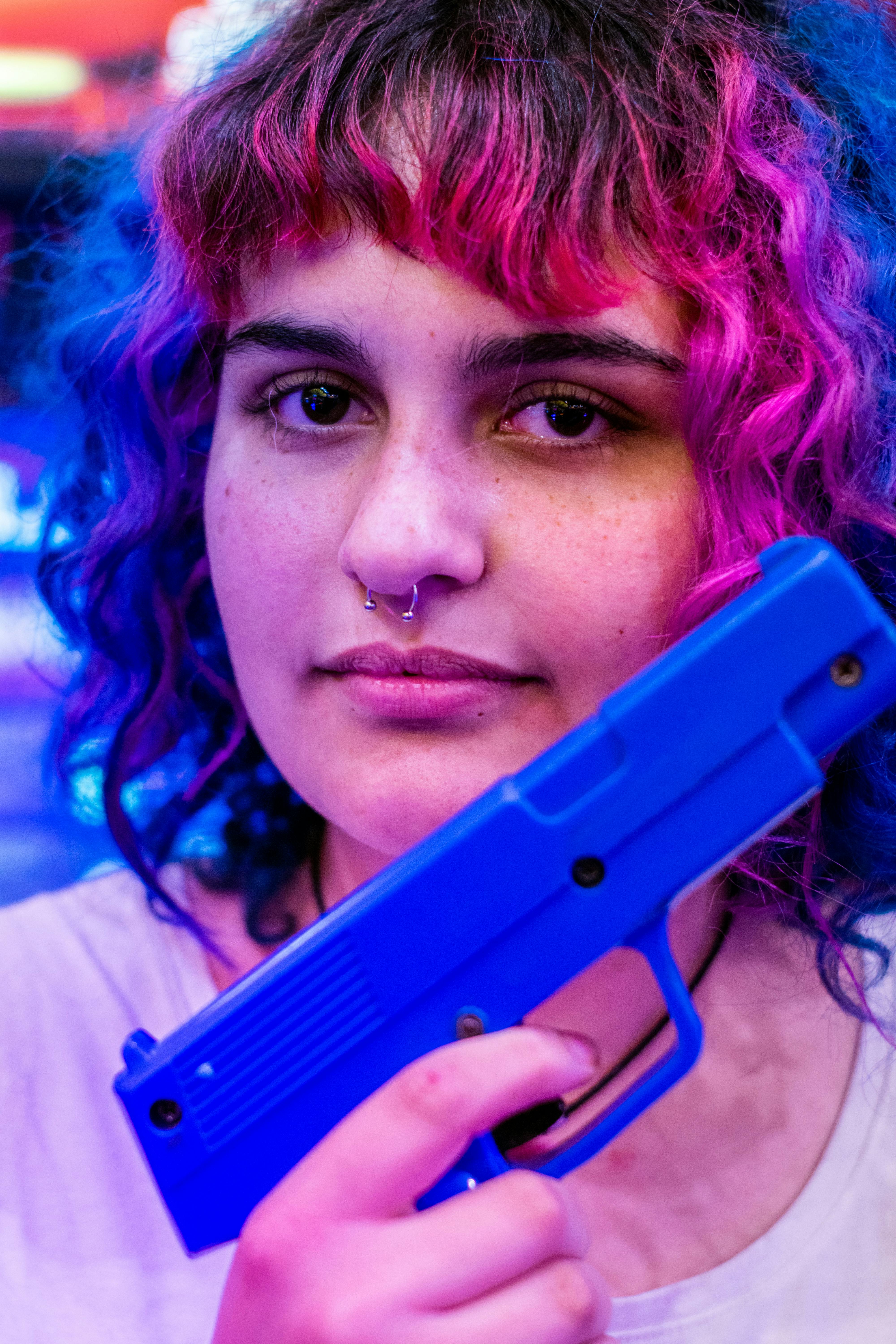 照片特写中，一名女子手持蓝色玩具枪，头发颜色鲜艳