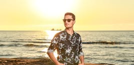 日落时分，一位穿着花衬衫、戴着太阳镜的男子在海滩边流露出自信