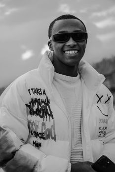 Black and white portrait of a fashionable young man smiling while wearing sunglasses and a graffiti jacket.