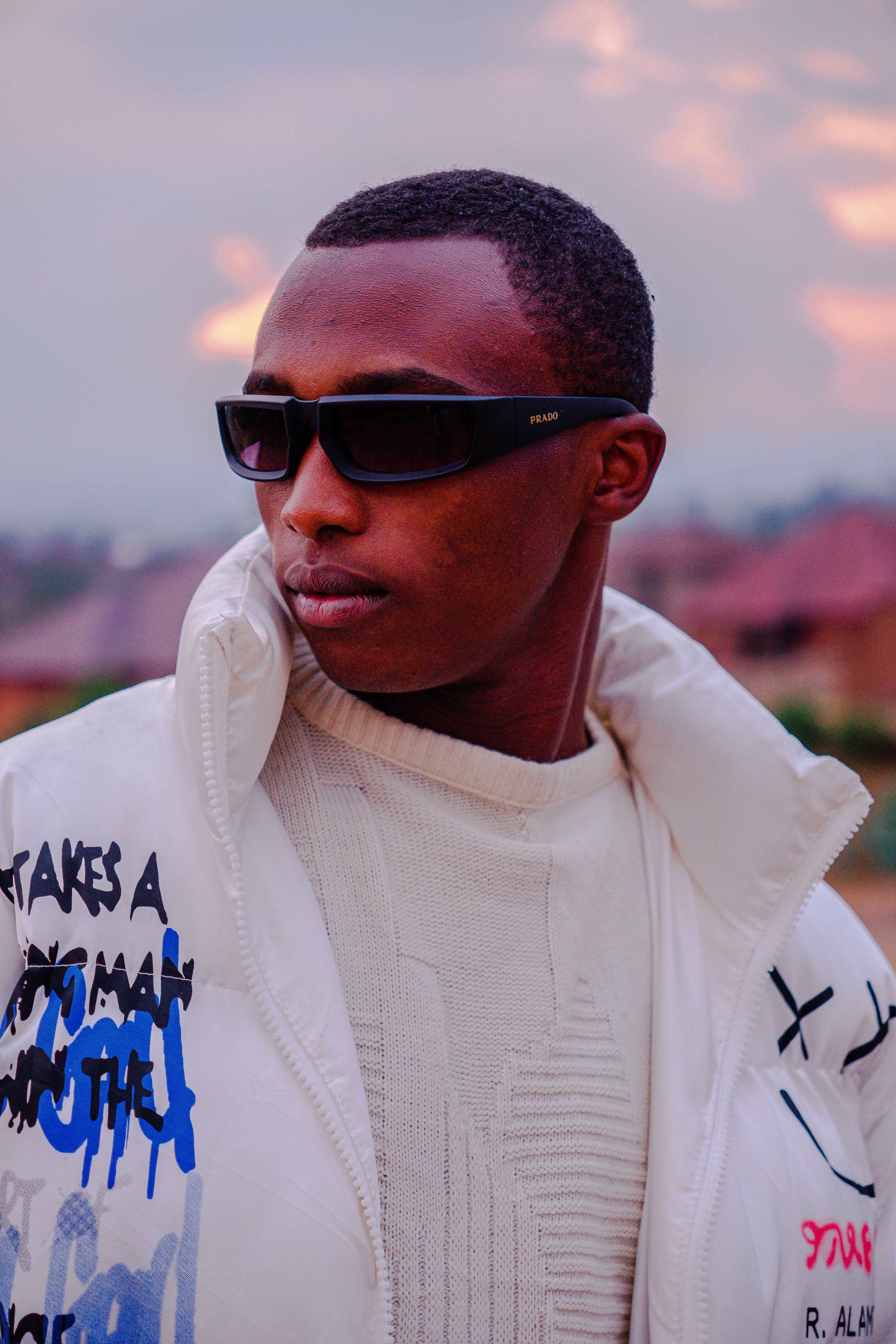Trendy man wearing stylish sunglasses and a white urban jacket outdoors during sunset.