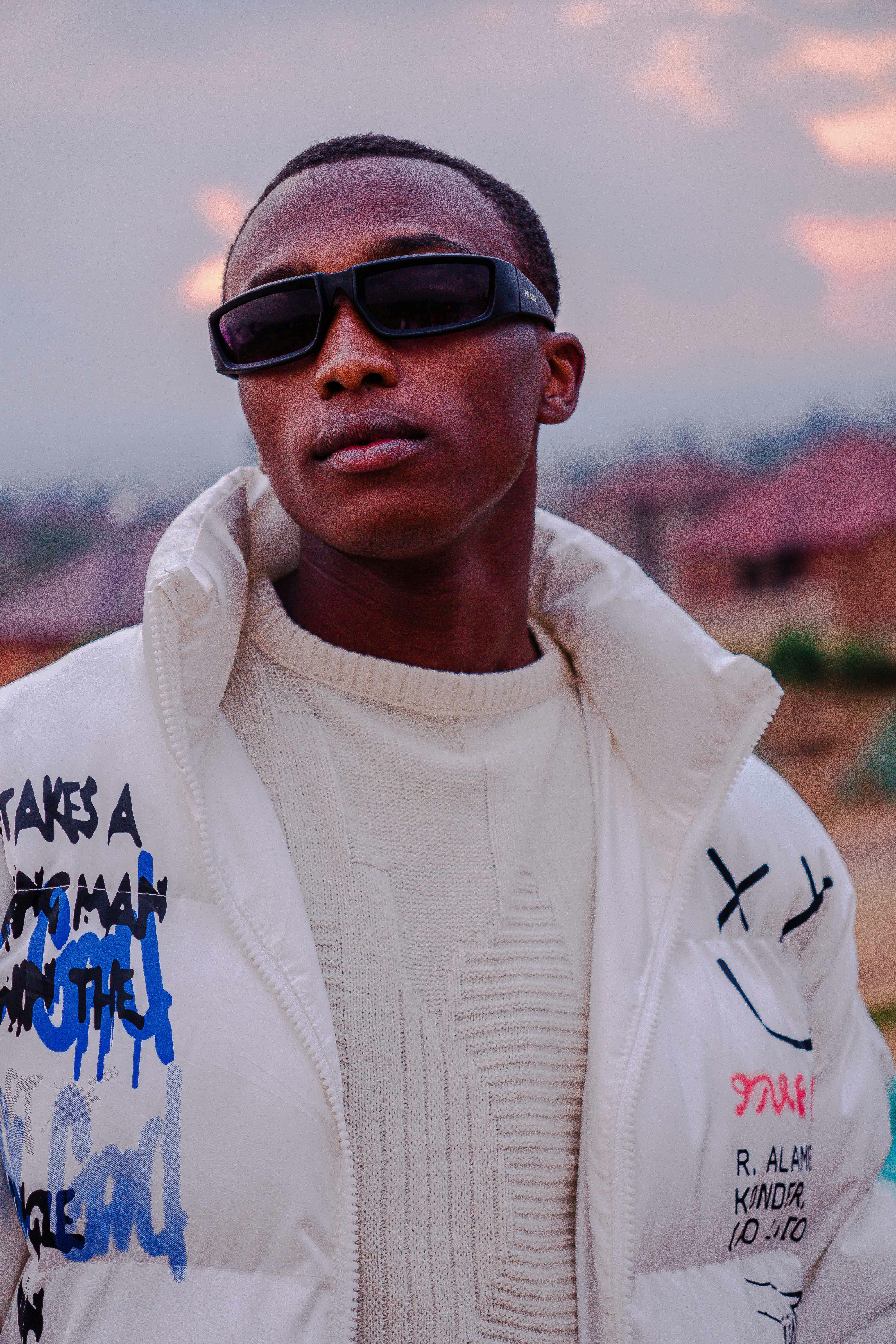 Stylish adult in a graphic jacket and sunglasses standing outdoors at sunset.