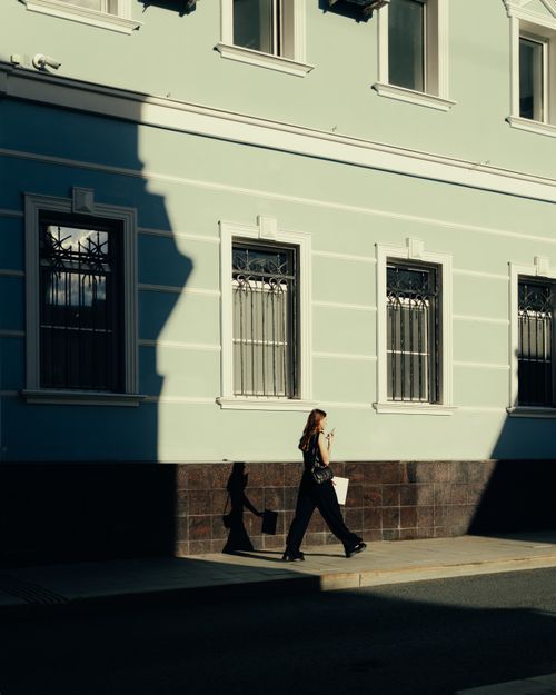 Free A woman walks along an urban street, casting dramatic shadows against a building. Stock Photo