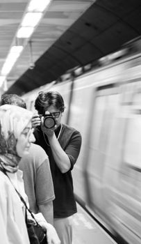 A photographer takes pictures in a busy subway station with a blurred train passing by.