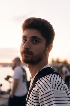 A trendy young man with facial hair in striped attire at a festival during sunset.