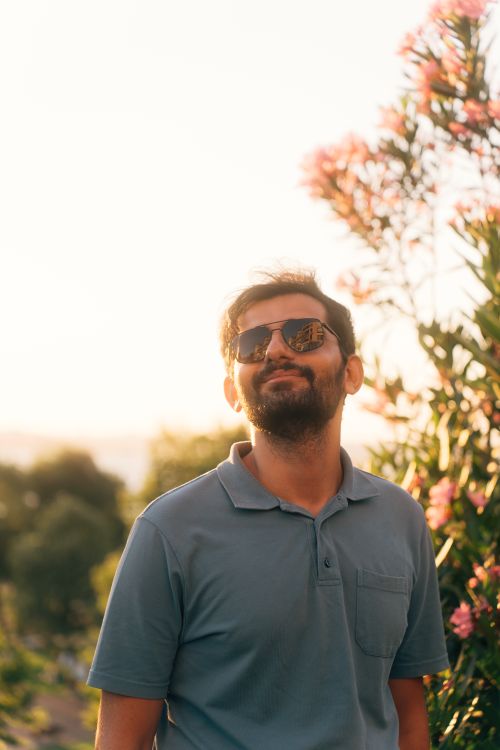 Free Portrait of a man wearing sunglasses, smiling outdoors with blooming flowers in summer light. Stock Photo