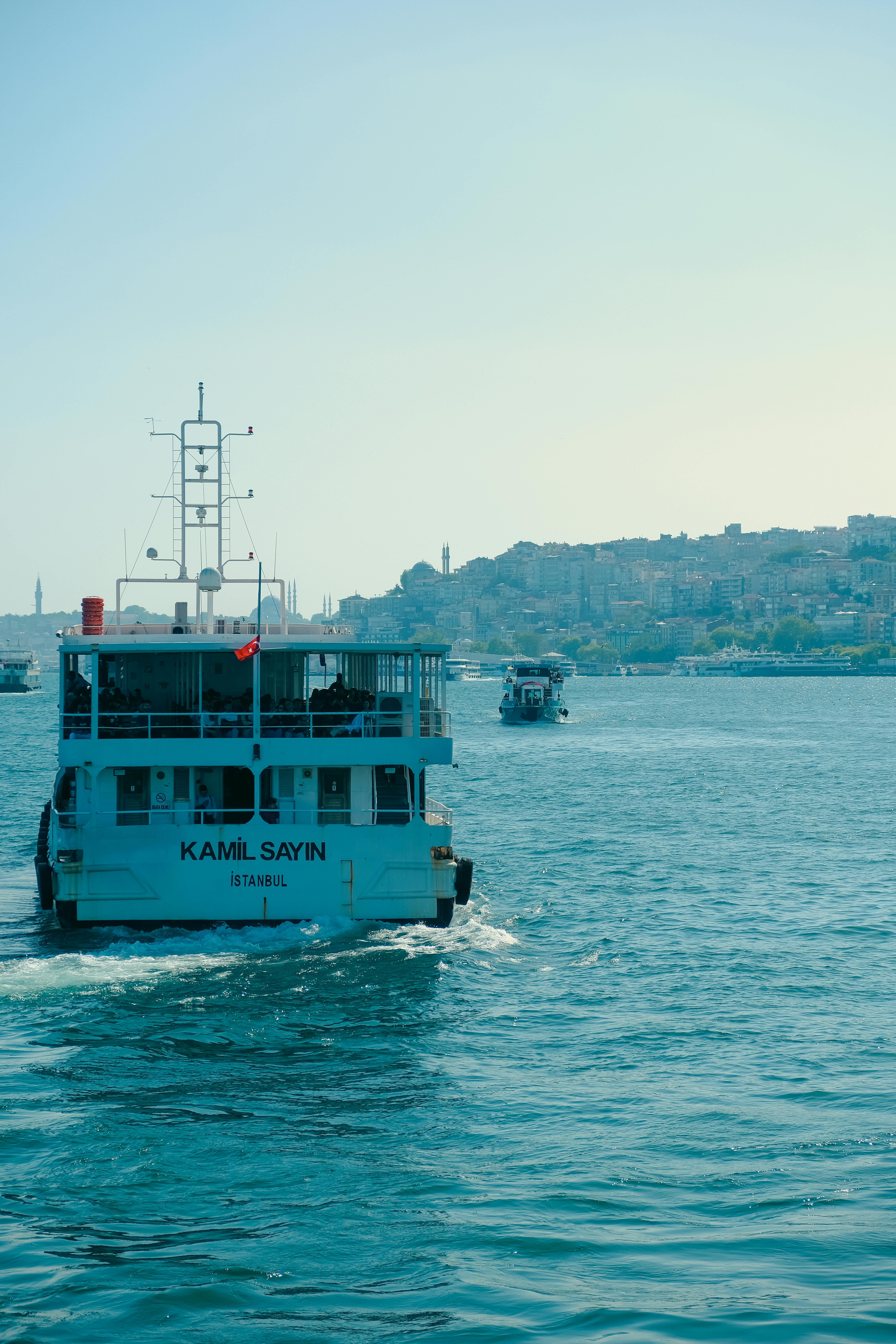 A ferry glides through Istanbul's waters with a cityscape backdrop under a clear summer sky.