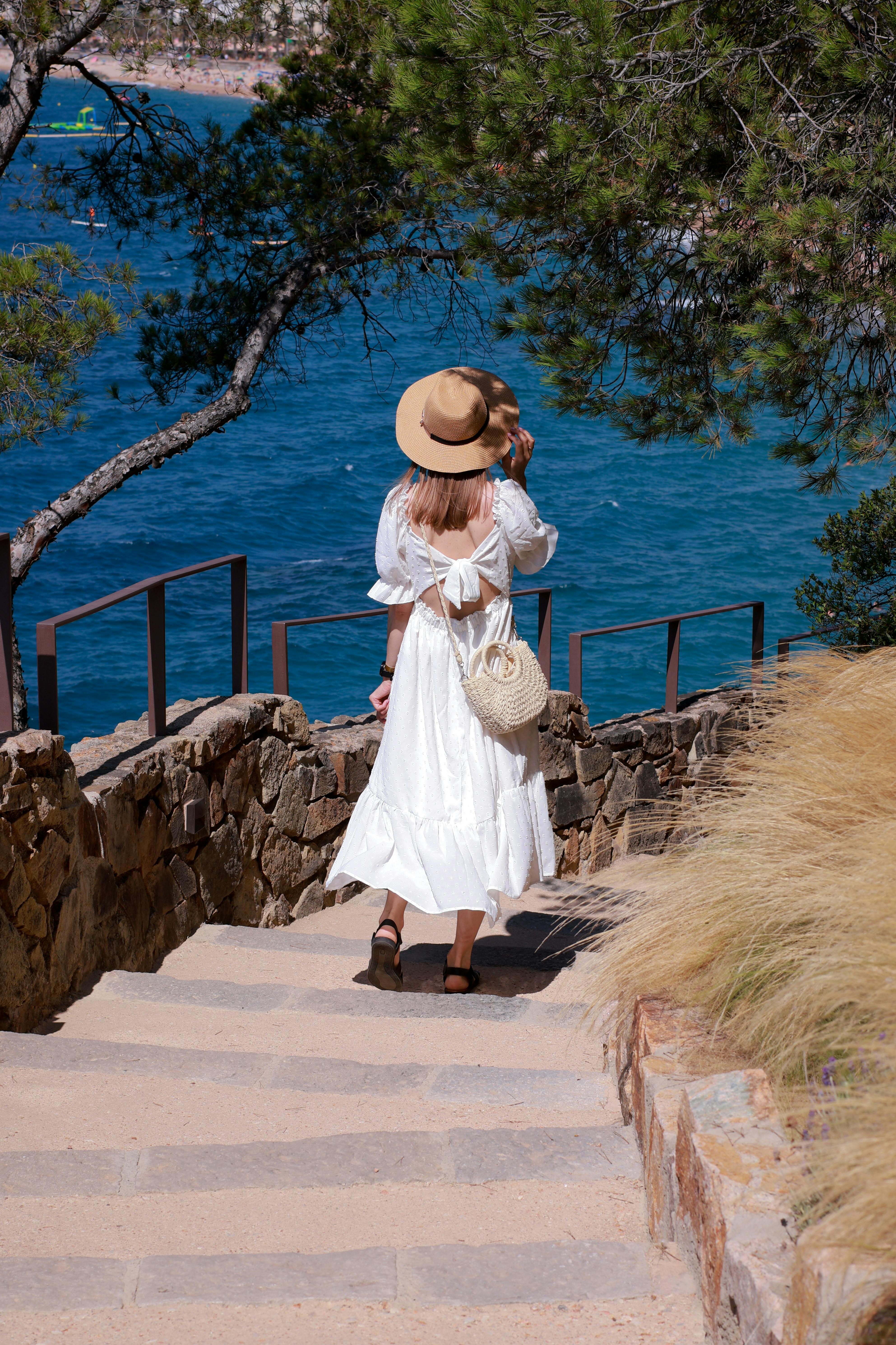 A woman in a white dress leisurely strolls down a stone path overlooking the blue sea on a sunny day.