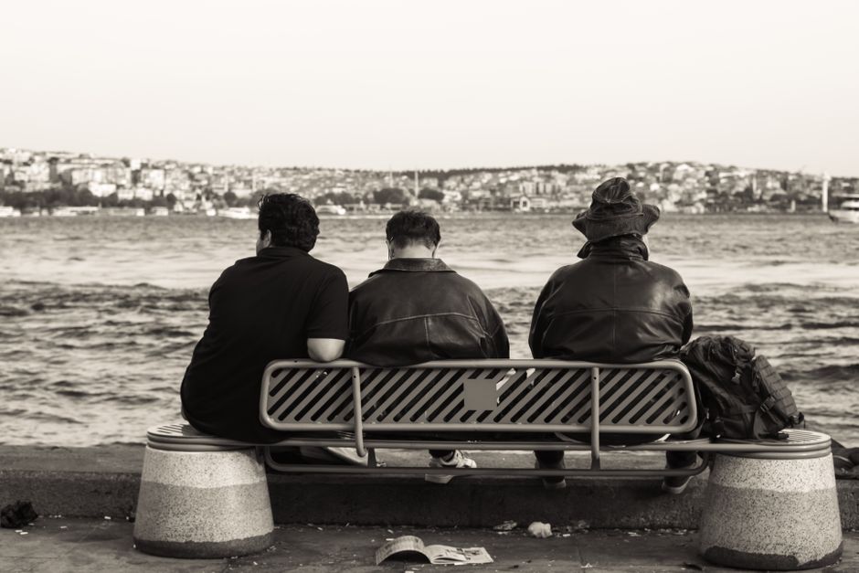 Three individuals sit on a waterfront bench, gazing across the water in monochrome style.