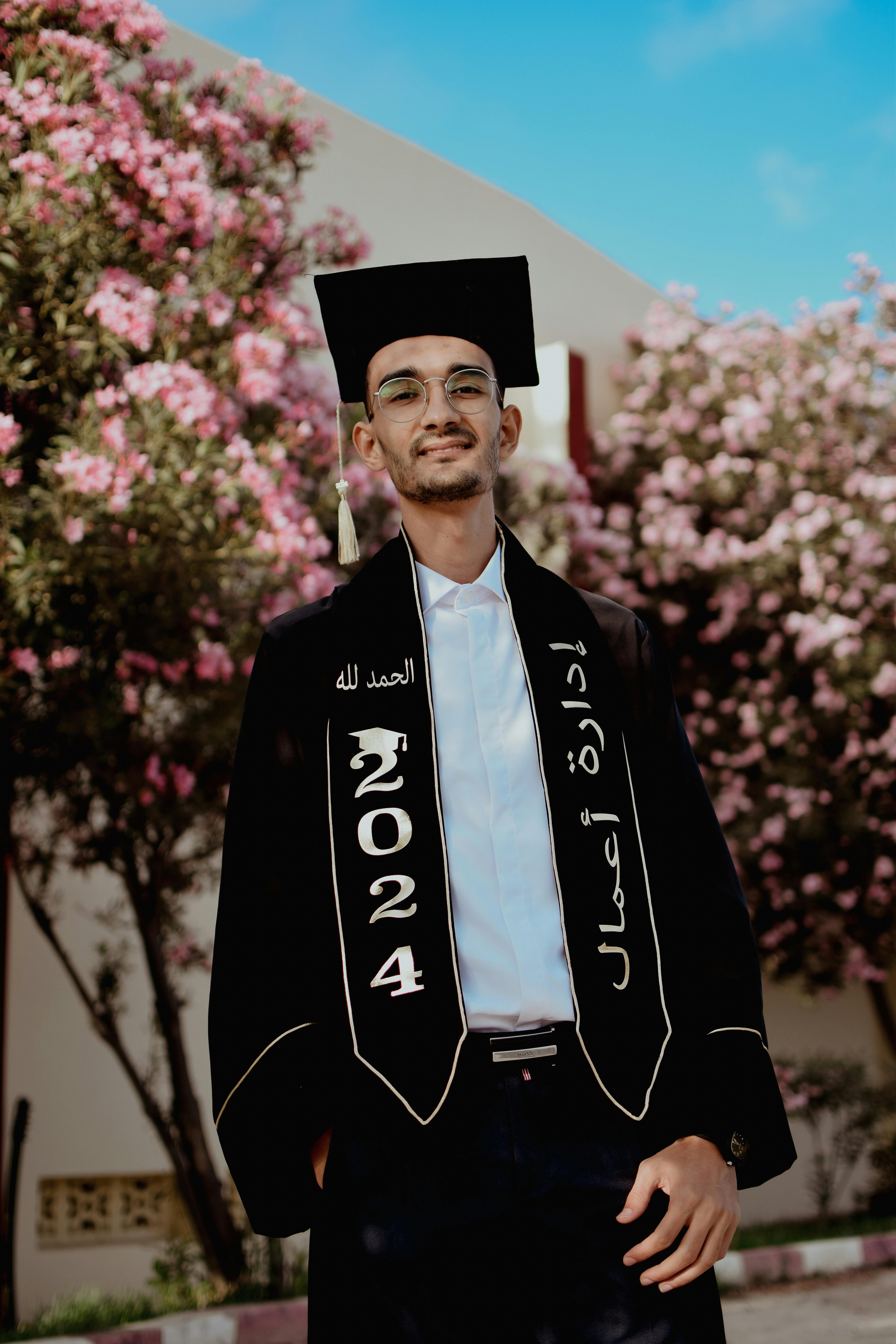 Confident young man in graduation attire stands proudly among blooming trees.