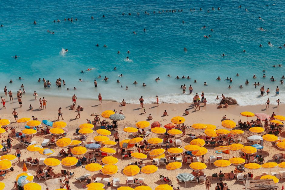 A vibrant beach scene in Kaş, Türkiye, with people enjoying the sea under yellow umbrellas.