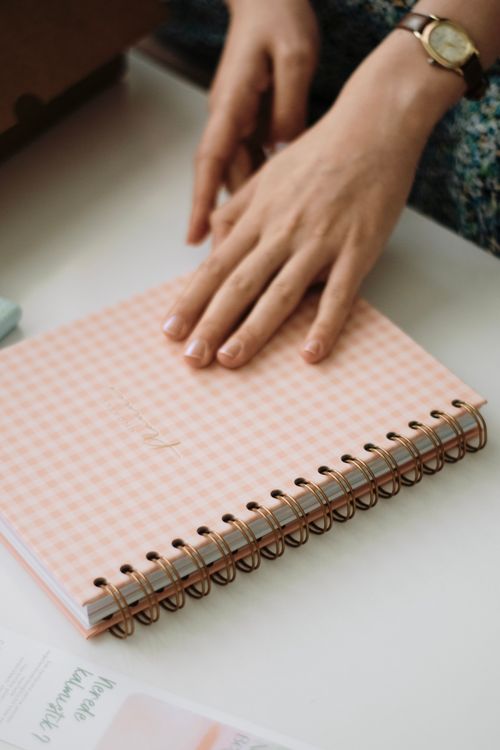 Free Close-up of a woman's hand resting on a spiral-bound notebook at a workspace. Stock Photo