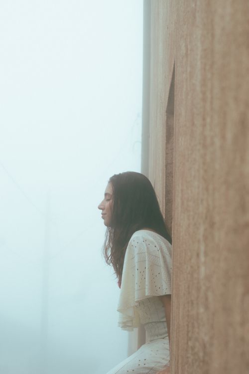 Free A woman in a white dress sits contemplatively by a wall, enveloped in fog. Stock Photo