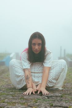 A young woman poses outdoors in a fog, wearing a white outfit and showcasing a tattoo.