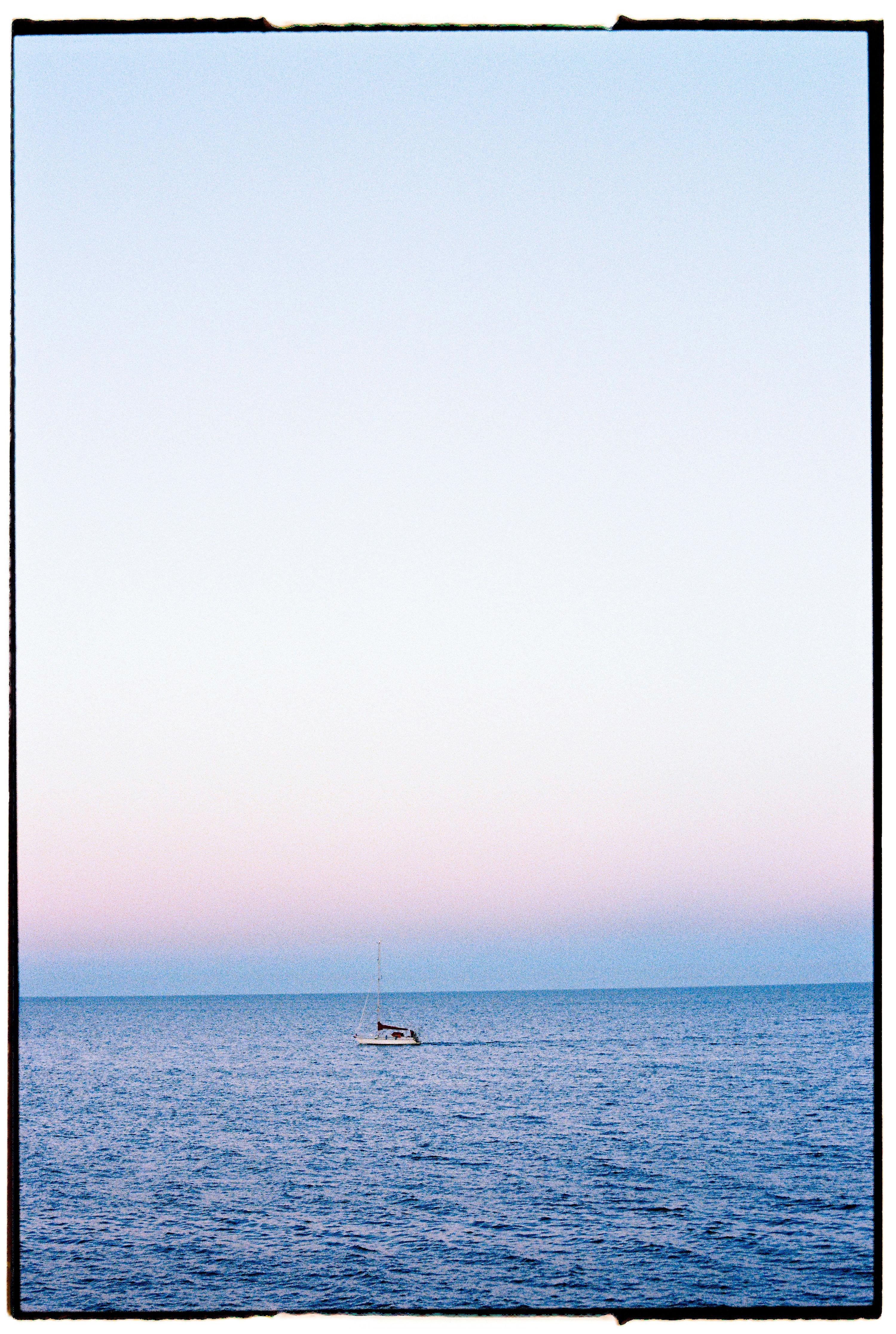 A lone sailboat glides across the calm ocean at dusk with a clear sky overhead.