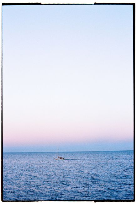 A lone sailboat glides across the calm ocean at dusk with a clear sky overhead.
