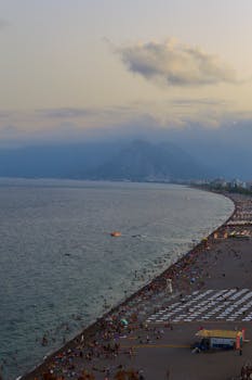 A scenic view of a bustling beach at sunset in Antalya, Turkey, with mountains and ocean.