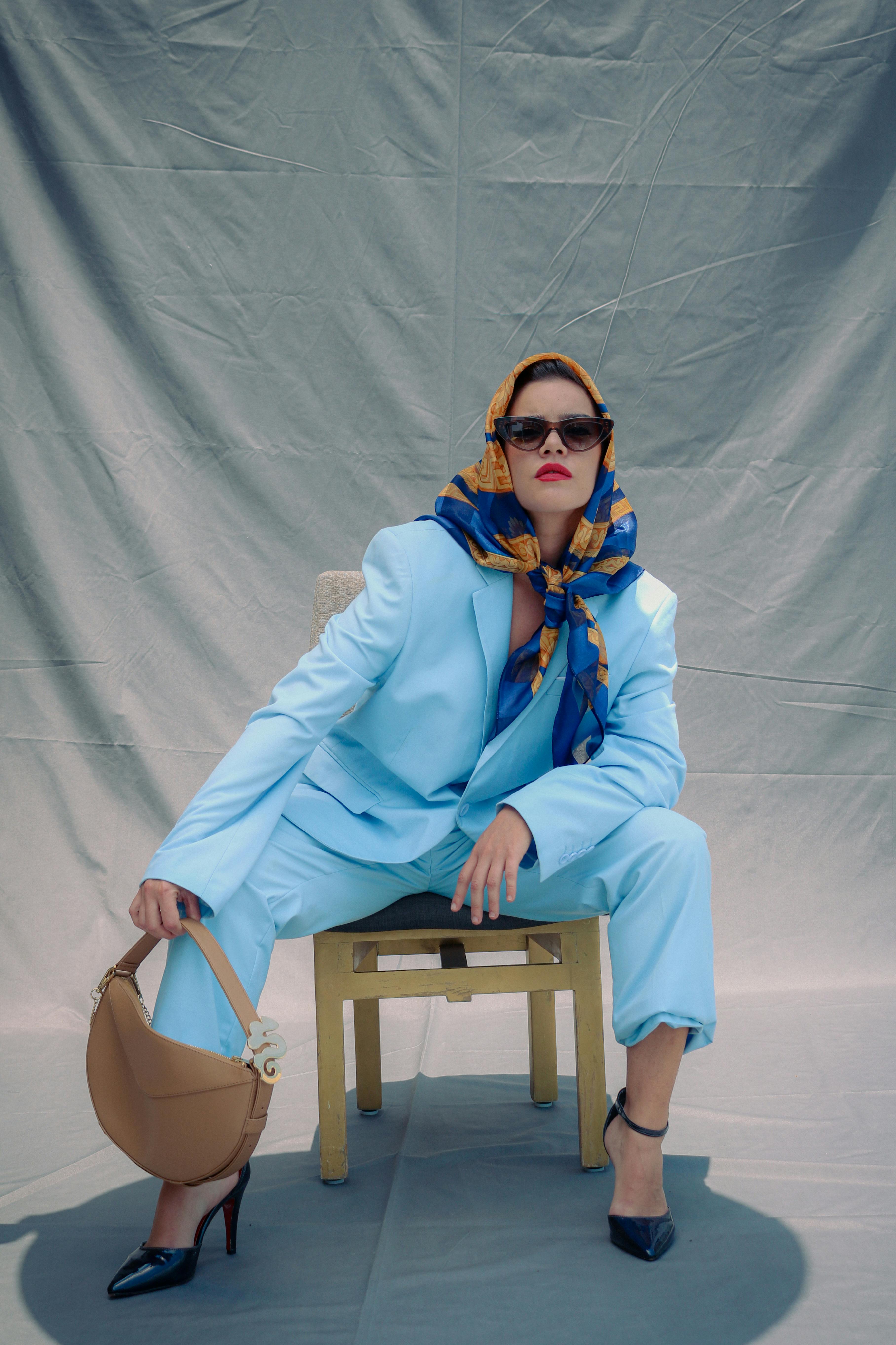 Stylish woman in blue suit and headscarf sitting on a chair in a studio setting.