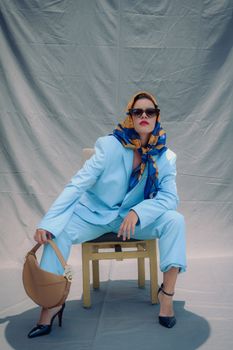 Stylish woman in blue suit and headscarf sitting on a chair in a studio setting.