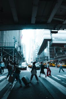 People crossing a busy street in New York City on a sunny day, featuring taxis and skyscrapers.