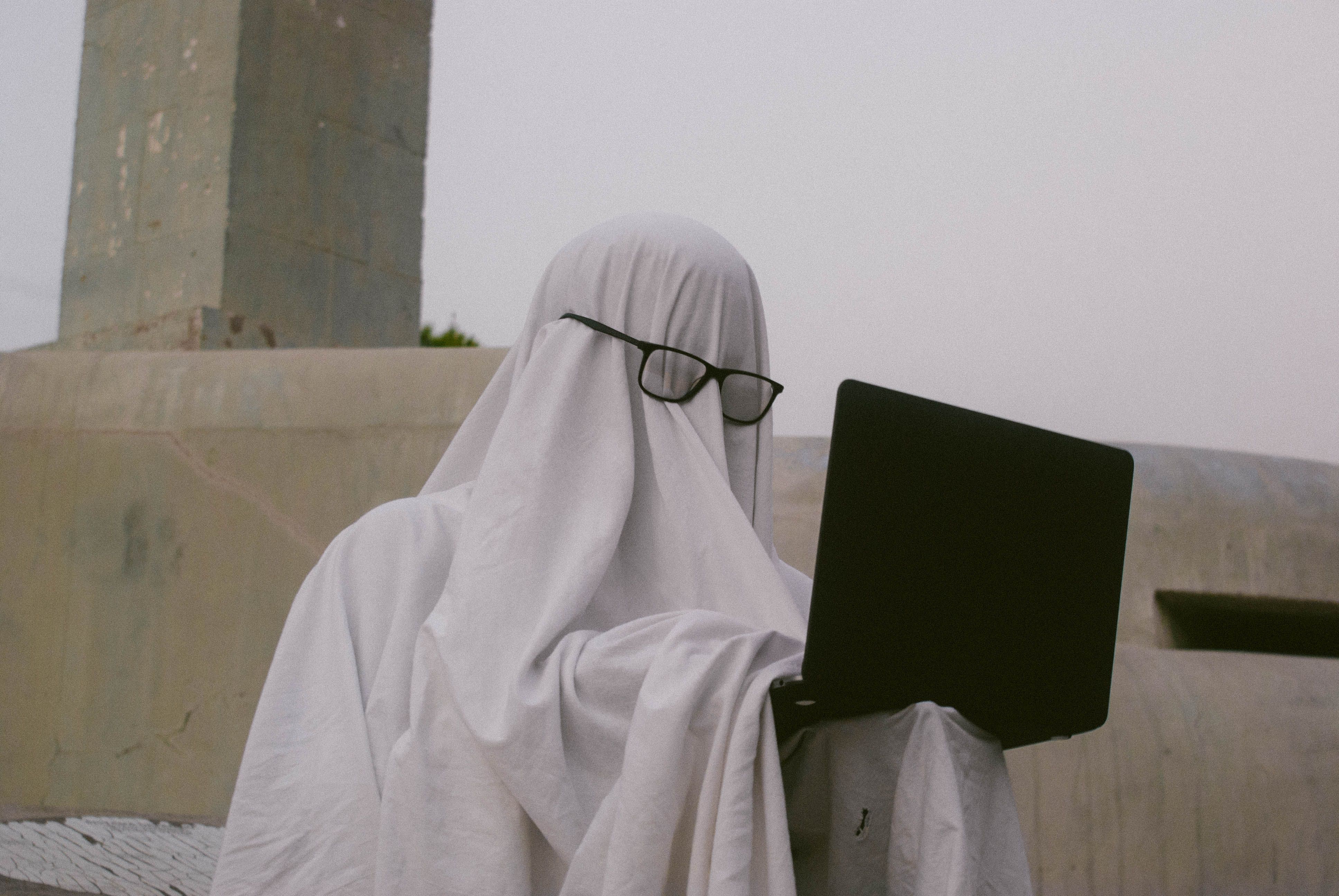 A humorous depiction of a person in a ghost costume using a laptop outdoors.