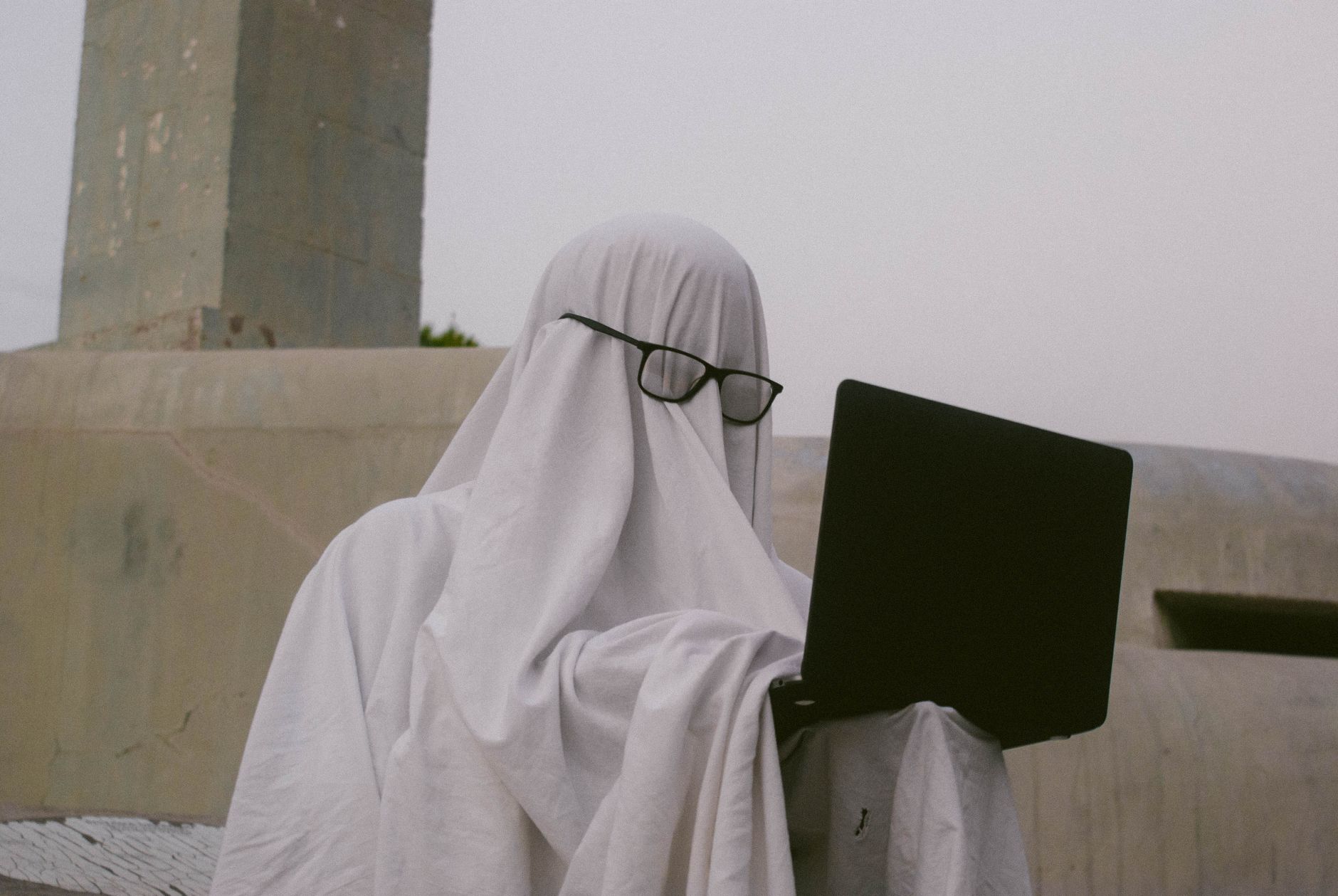 A humorous depiction of a person in a ghost costume using a laptop outdoors.