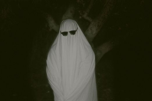 Mysterious figure dressed as a ghost wearing sunglasses in a dark forest setting.