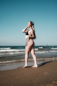 A woman in a bikini poses confidently by the ocean on a sunny day.