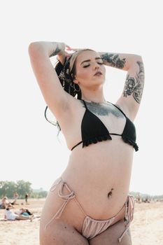 Confident woman in a black bikini with tattoos posing on a sunny beach, exuding a powerful and fashionable vibe.