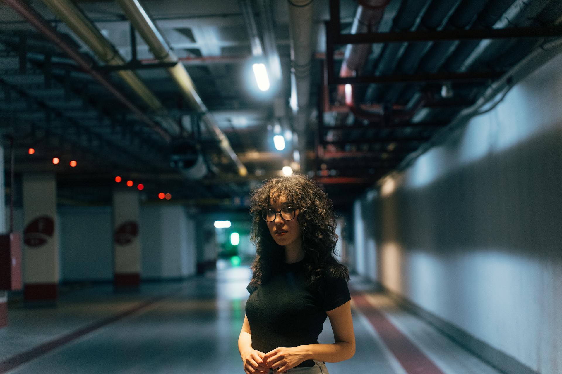 A woman with curly hair stands in an urban tunnel, surrounded by industrial pipes and lighting.