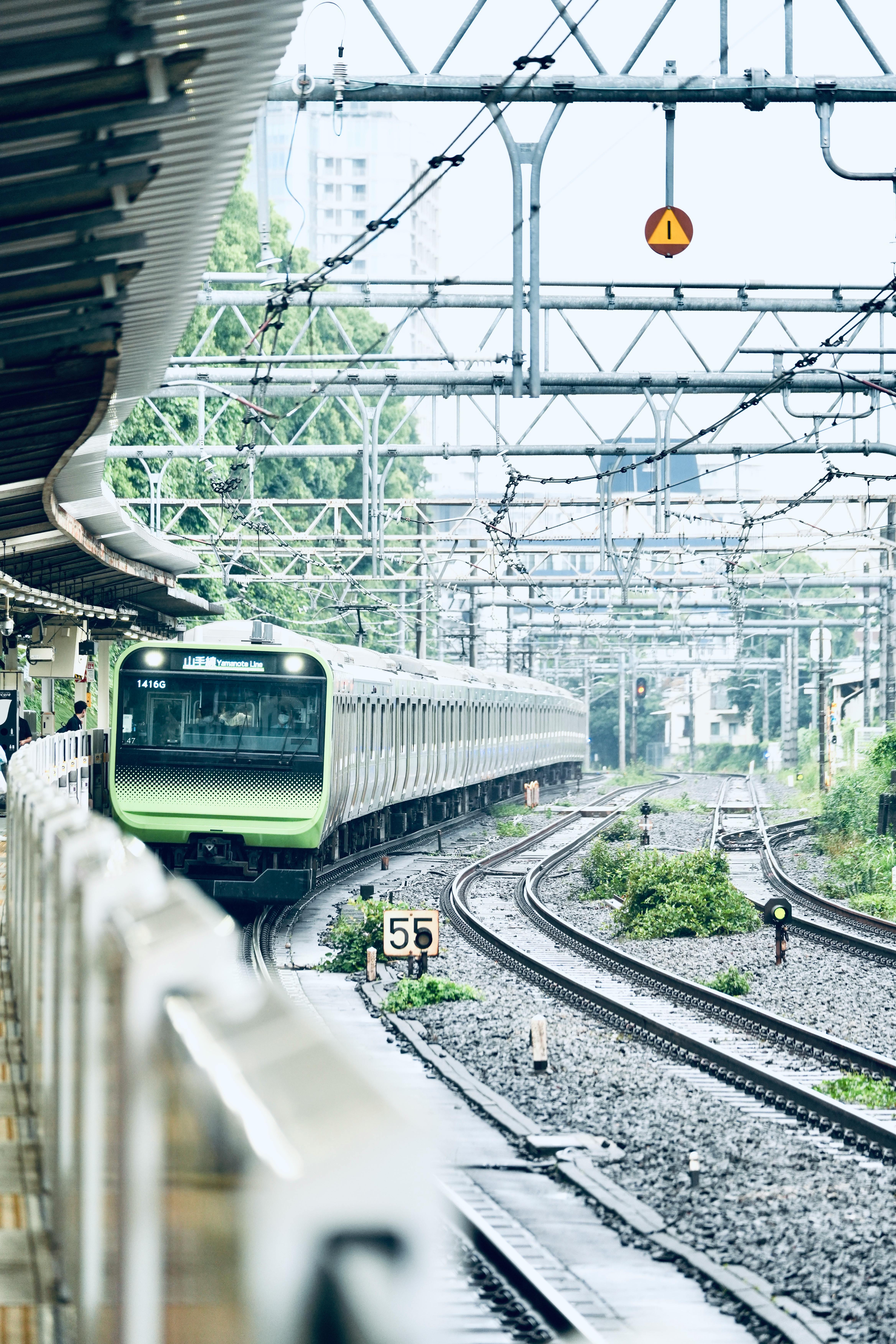 A sleek green train approaching a city station under electric lines on a cloudy day.