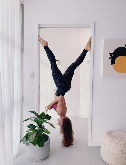 Free Woman in Workout Gear Hanging Upside Down on Doorway Near Green Plants Stock Photo