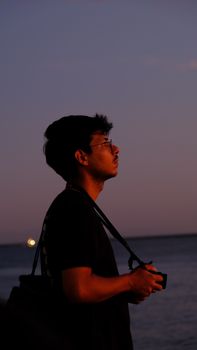 A man with a camera looks thoughtfully towards the ocean at sunset.