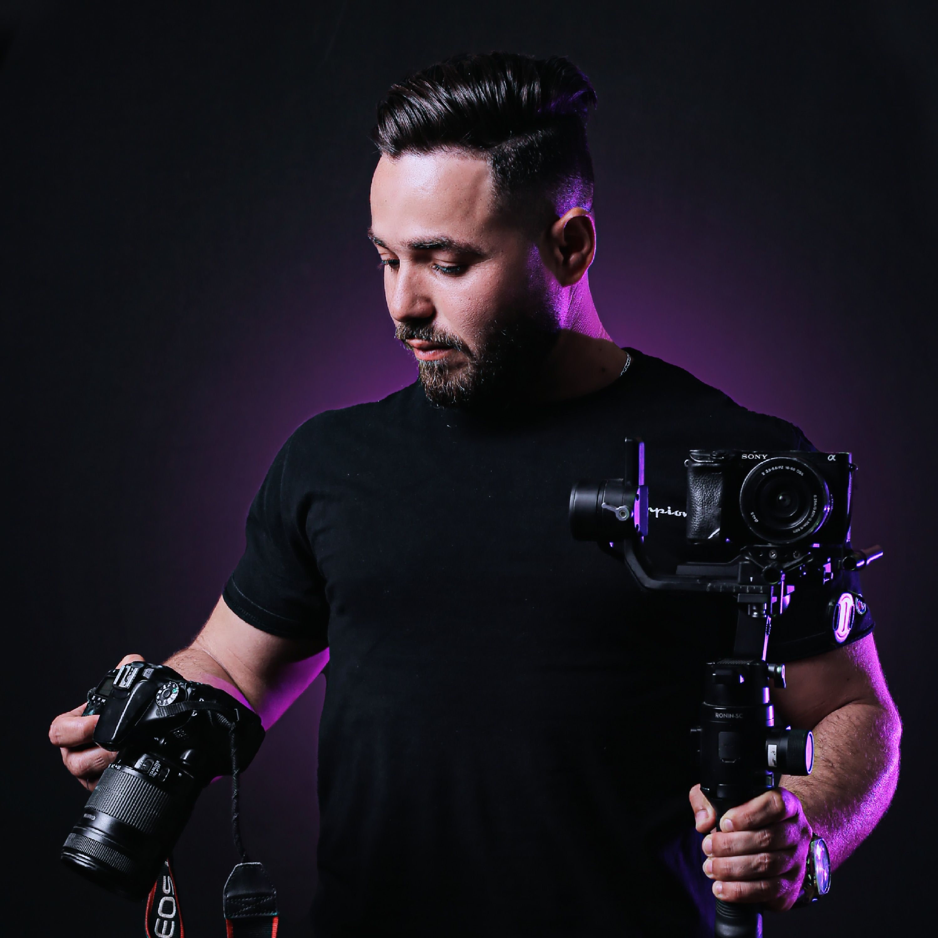 Portrait of a professional photographer holding cameras in a studio with purple ambient lighting.